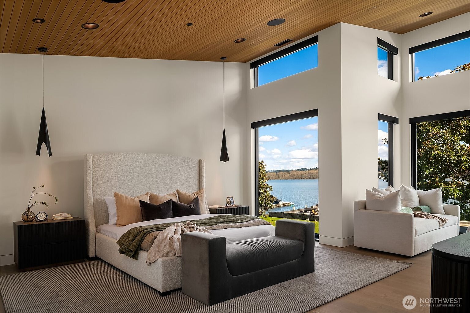 This luxurious primary bedroom features a high, wood-paneled vaulted ceiling and expansive floor-to-ceiling windows that offer a stunning, unobstructed view of the lake. The room is anchored by a plush, textured upholstered bed and a sleek dark wood bench, complemented by a cozy seating area and minimalist pendant lighting. The overall aesthetic is modern, serene, and sophisticated, emphasizing the seamless connection between the interior living space and the natural waterfront surroundings.