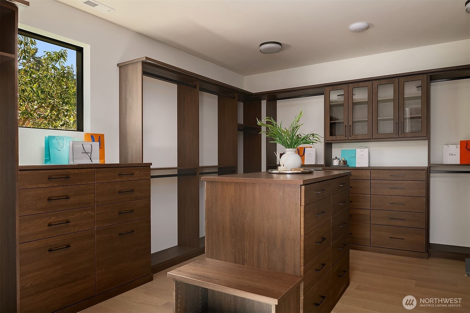 This spacious, high-end walk-in closet features custom dark wood cabinetry, a central island with multiple drawers, and ample hanging space. The room is accented with a window providing natural light, decorative shopping bags, and a small plant, creating a luxurious and organized atmosphere. The perspective is a wide-angle shot from the entrance, showcasing the functional layout and sophisticated design.