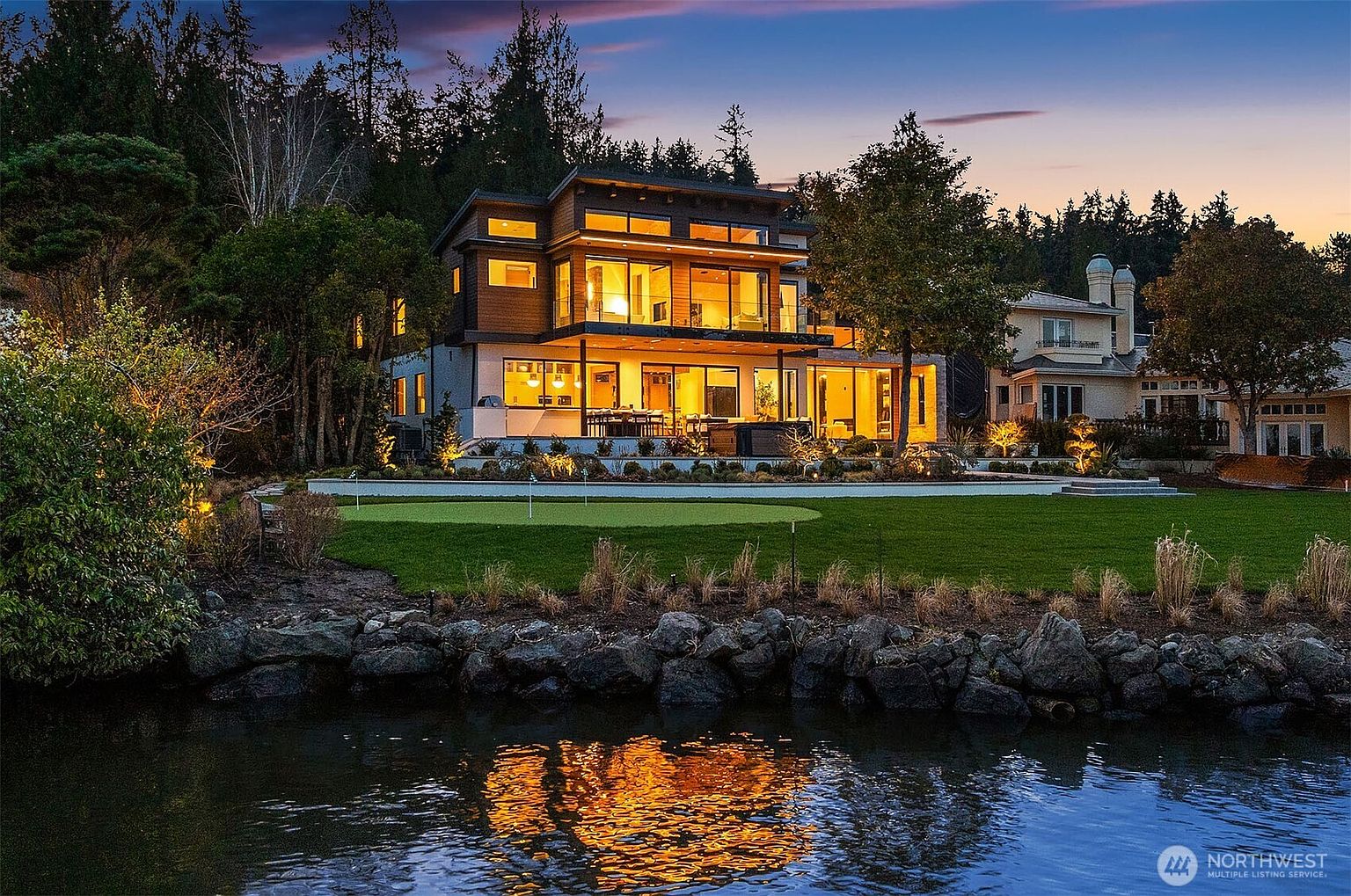 This stunning contemporary waterfront home is captured at dusk, showcasing its expansive glass-walled design and warm, inviting interior lighting. The property features a meticulously manicured lawn with a private putting green, leading directly to a rocky shoreline. The cinematic perspective highlights the seamless transition between the modern architecture and the serene natural landscape.