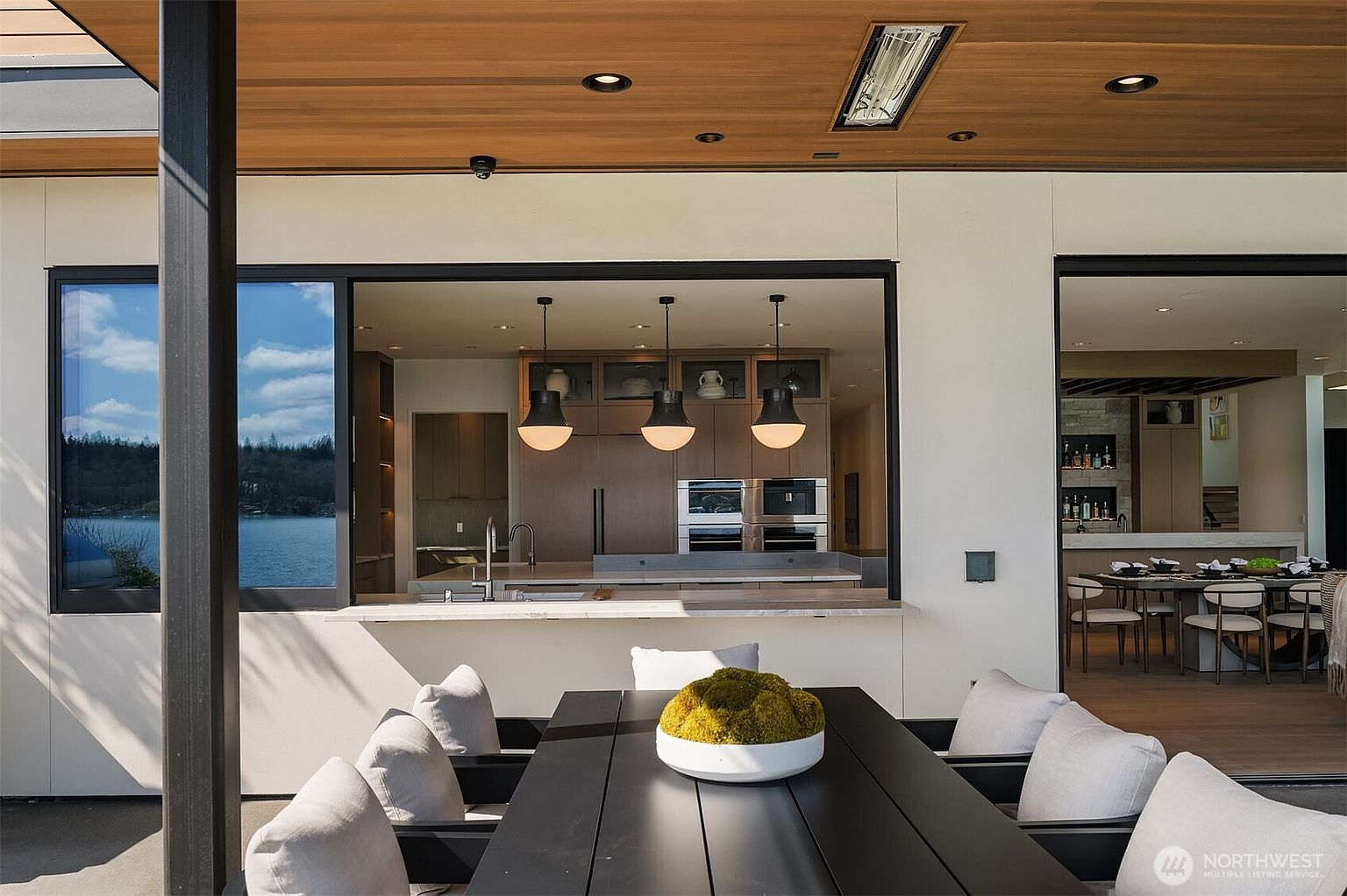 This sophisticated outdoor patio space features a sleek, modern dining set positioned under a covered ceiling with recessed lighting and a built-in heater. The area seamlessly connects to an indoor kitchen through a large pass-through window, creating an ideal layout for entertaining while offering a scenic view of the water. The design emphasizes a clean, contemporary aesthetic with high-end finishes and a smooth transition between indoor and outdoor living.