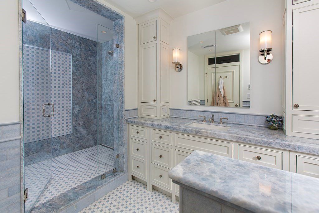 This is a well-lit primary bathroom featuring a glass-enclosed shower with blue-patterned tile, a white vanity with a blue marble countertop, and a large mirror with sconces. The bathroom has a clean and elegant design with a cohesive color scheme, creating a relaxing and luxurious atmosphere. The floor tiles match the shower tiles, enhancing the visual harmony.