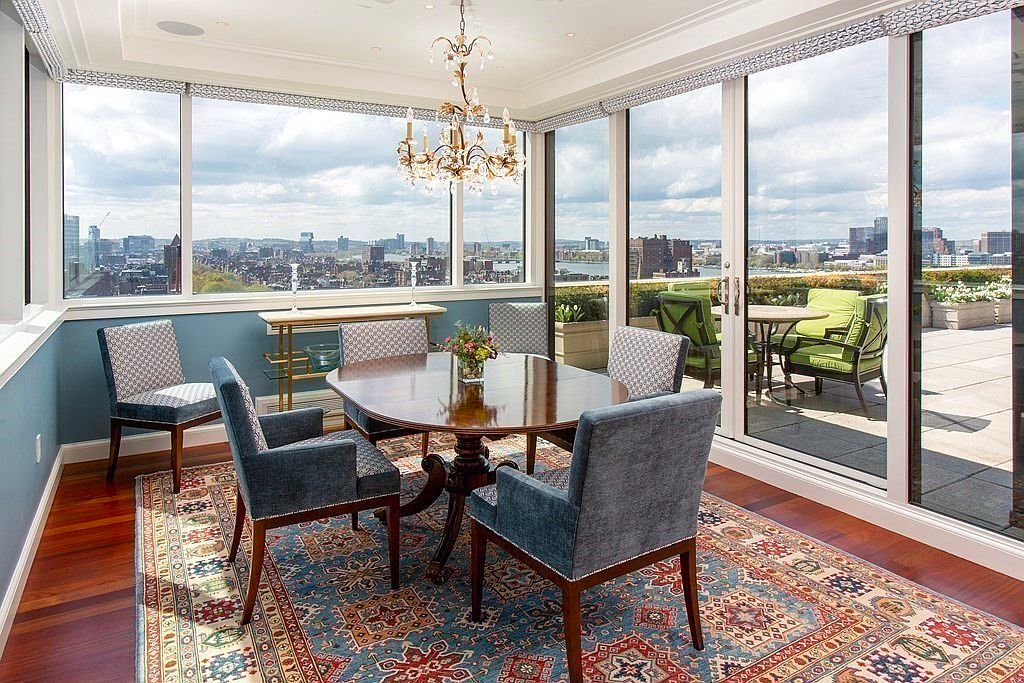 This is an interior shot of a dining room featuring a round wooden table surrounded by upholstered chairs. Large windows offer a panoramic city view, and a chandelier hangs above the table. The room is decorated with a patterned rug and a small console table, creating an elegant and inviting atmosphere.