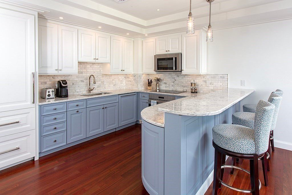 This is a well-lit kitchen featuring two-tone cabinetry, with white upper cabinets and light blue lower cabinets. The countertops are a light-colored granite, and the backsplash is a marble tile. The kitchen also features a breakfast bar with three stools and stainless steel appliances, creating a modern and inviting space.