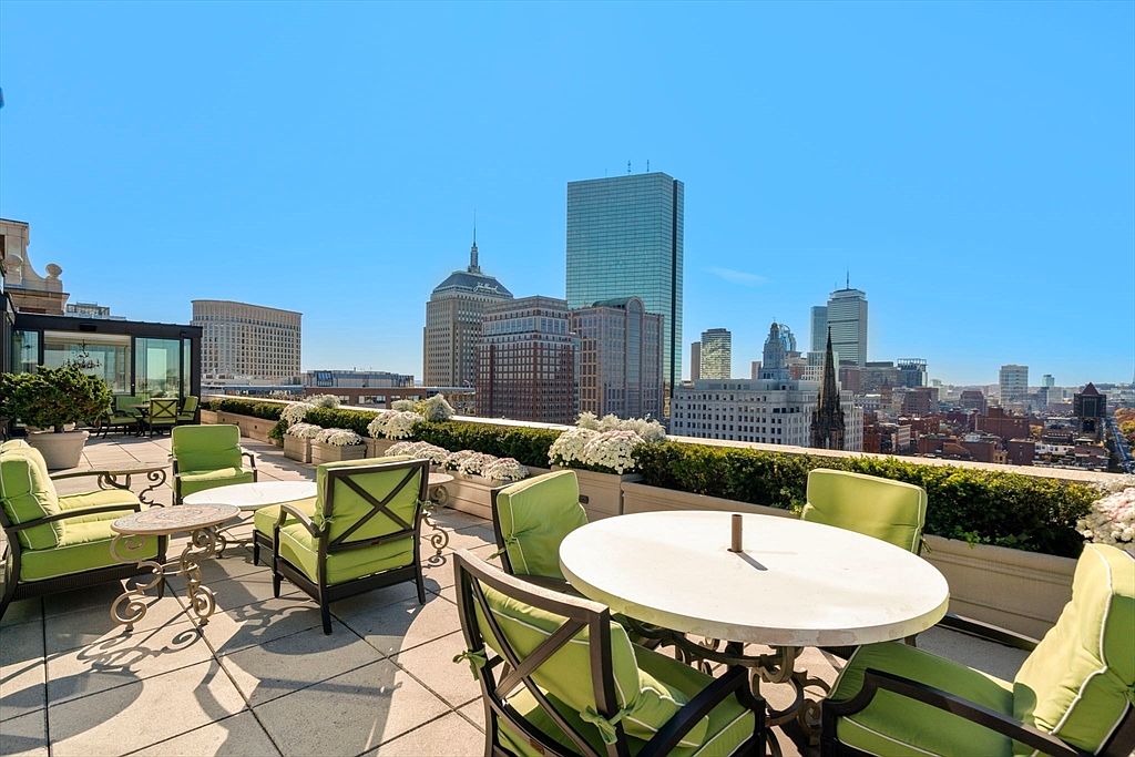 This image showcases a luxurious rooftop patio or balcony with stunning city views. The space is furnished with comfortable seating arrangements, including cushioned chairs and round tables, surrounded by lush greenery and flower boxes. The overall impression is one of upscale urban living and relaxation.