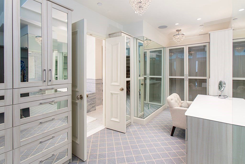 This is a well-organized walk-in closet featuring mirrored cabinets and drawers, providing ample storage space. The room is illuminated by elegant chandeliers, and the patterned carpet adds a touch of sophistication. An open doorway leads to an adjacent bathroom, enhancing the convenience and luxury of the space.