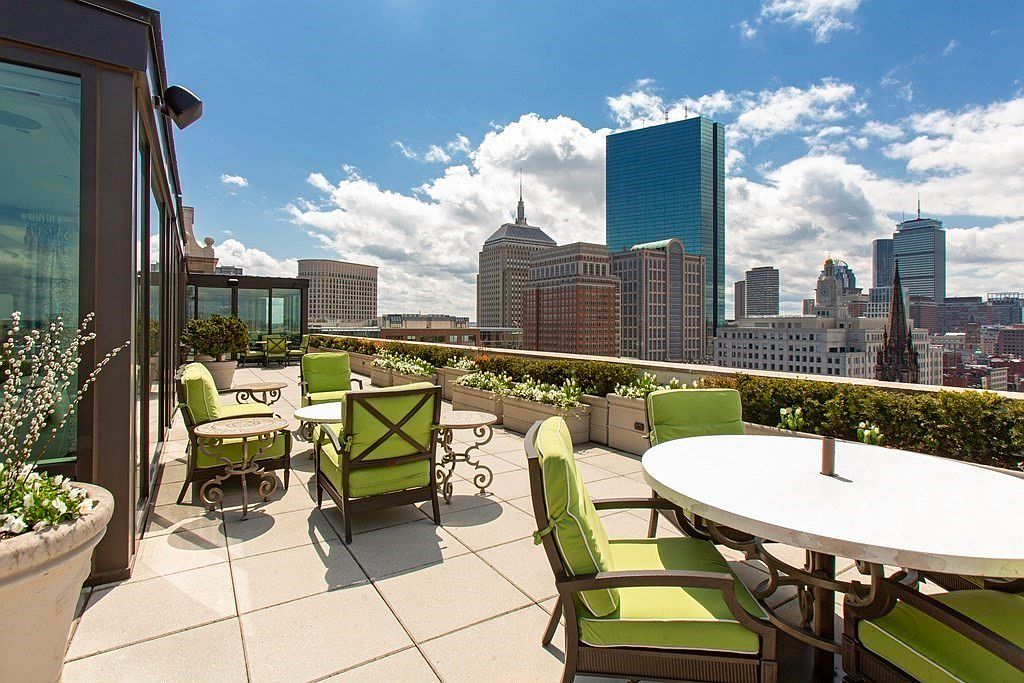 This image showcases a luxurious rooftop patio or balcony with stunning city views. The space is furnished with comfortable seating arrangements, including chairs and a round table, all featuring green cushions. Lush greenery lines the perimeter, adding a touch of nature to the urban setting, while the cityscape provides a dramatic backdrop.