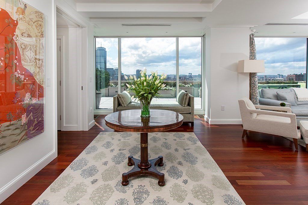 This interior shot showcases a sophisticated living room with hardwood floors and a decorative area rug. A round table with a floral arrangement sits in the foreground, leading to a seating area with a daybed and a sofa, all set against a backdrop of large windows offering city views. The room exudes an elegant and comfortable atmosphere, perfect for relaxation and entertaining.