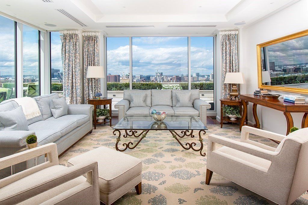This is a bright and elegant living room featuring large windows with city views. The room is furnished with two sofas, two armchairs, a glass-topped coffee table, and a wooden console table with a gold-framed mirror above it. The decor is sophisticated and inviting, creating a luxurious and comfortable atmosphere.
