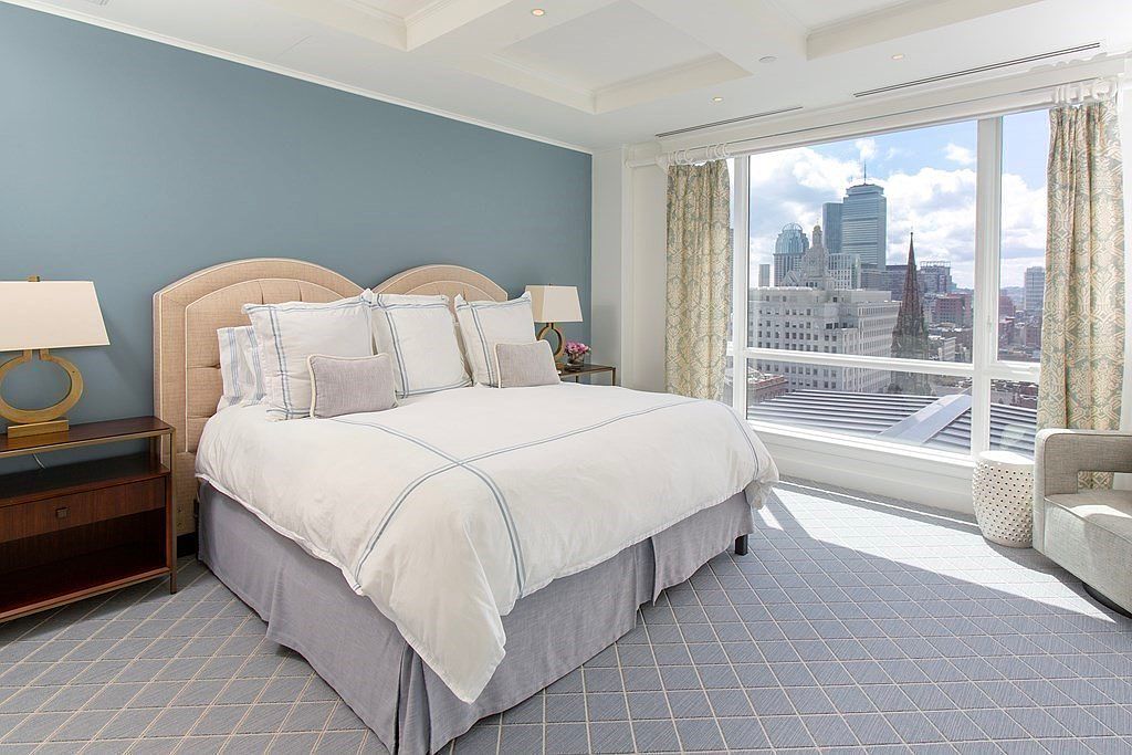 This primary bedroom features a large bed with a neutral-toned headboard and white linens, complemented by a blue accent wall. A large window offers a stunning city view, enhancing the room's spacious and luxurious feel. The room is well-lit and decorated in a modern style.