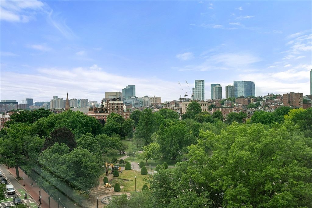 This aerial view showcases a vibrant cityscape with lush green trees in the foreground and a skyline of modern buildings in the background. The image captures a blend of urban and natural elements, suggesting a desirable location with access to both city amenities and green spaces. The clear blue sky enhances the overall appeal, making it an attractive prospect for potential buyers.