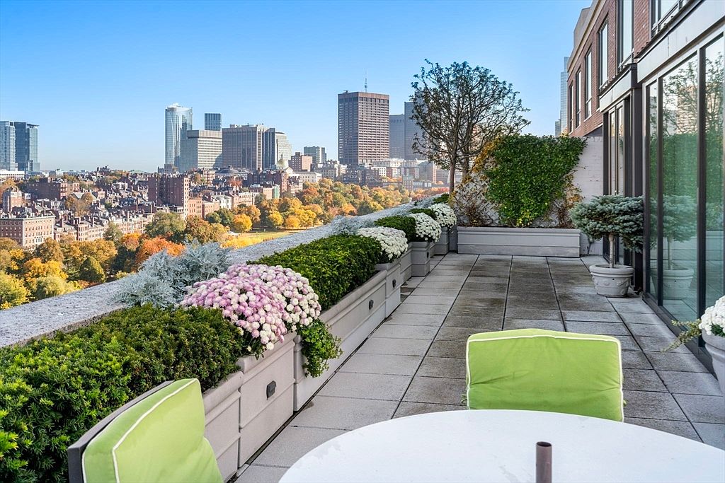 This image showcases a luxurious rooftop balcony with stunning city views. The balcony features a tiled floor, lush greenery in planters along the edge, and comfortable outdoor seating. The overall impression is one of sophisticated urban living with a private outdoor space.