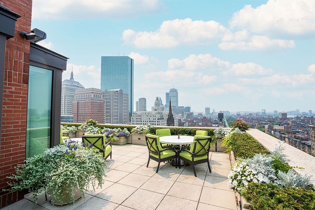 This image showcases a luxurious rooftop patio with stunning city views. The patio features a tiled surface, comfortable outdoor seating with green cushions around a white table, and lush planters filled with greenery and flowers. The backdrop includes a skyline with prominent skyscrapers under a partly cloudy sky, creating an inviting and upscale outdoor living space.