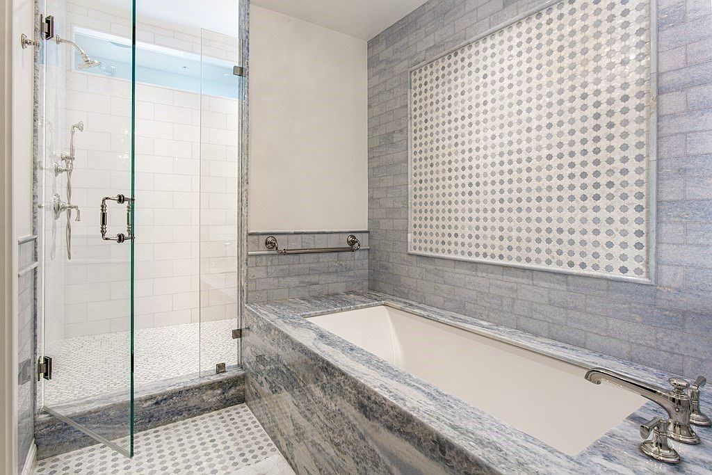 This is a luxurious primary bathroom featuring a glass-enclosed shower with white subway tile and a pebble floor. A soaking tub is surrounded by blue marble and accented with a decorative tile panel. The overall impression is clean, modern, and spa-like.
