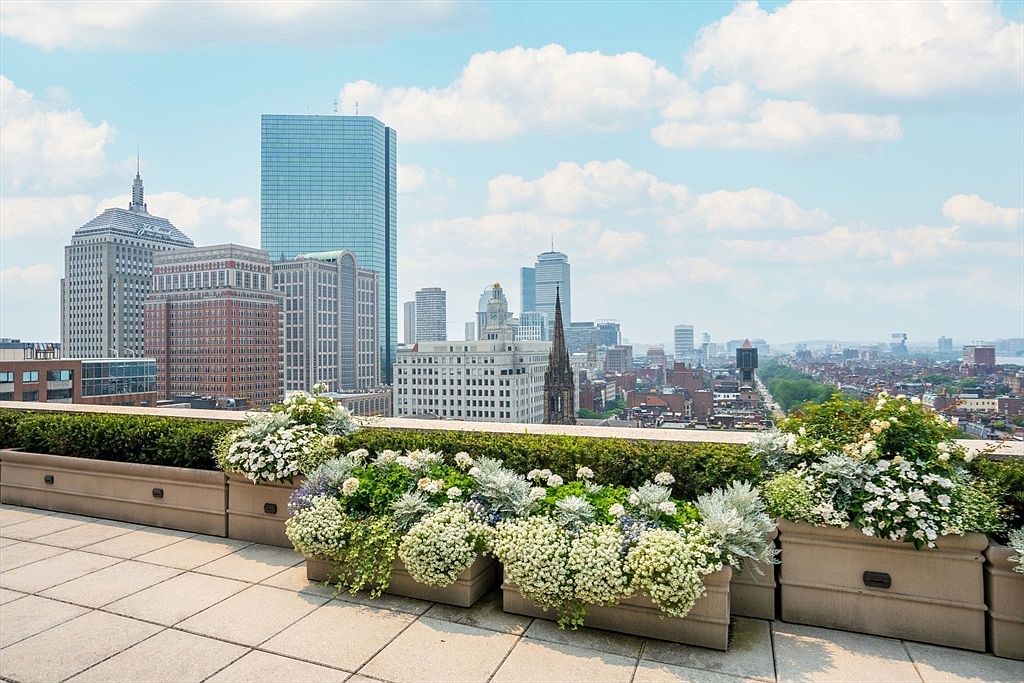 This image showcases a luxurious patio or balcony with a stunning cityscape view. The foreground features well-maintained planters filled with lush greenery and white flowers, adding a touch of nature to the urban setting. The background reveals a skyline with prominent skyscrapers under a partly cloudy sky, creating a sense of grandeur and sophistication.