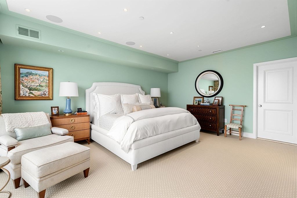 This is a primary bedroom featuring a large white bed with white linens, a wooden nightstand with a blue lamp, and a comfortable armchair with an ottoman. The walls are painted a soft green, and the room is well-lit with recessed lighting. A dark wood dresser with a round mirror sits against the far wall, adding a touch of elegance.