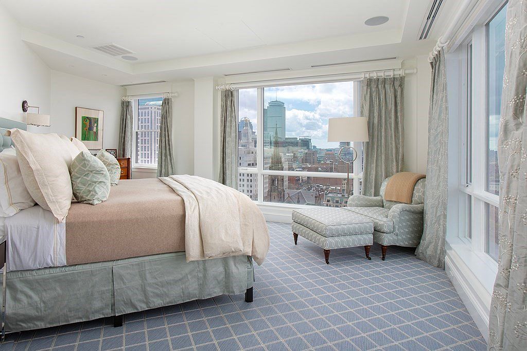 This is a primary bedroom featuring a large bed with neutral-toned bedding and decorative pillows. The room has large windows offering a city view, complemented by light-colored curtains. An armchair and ottoman are positioned near the window, and the room is carpeted with a geometric pattern, creating a serene and luxurious atmosphere.