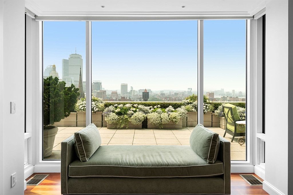 This sunroom features a comfortable daybed positioned in front of large windows that offer a panoramic city view. The room is bright and airy, with hardwood floors and white trim, creating a relaxing and inviting space. The outdoor terrace is visible through the windows, adorned with lush greenery and flowers.