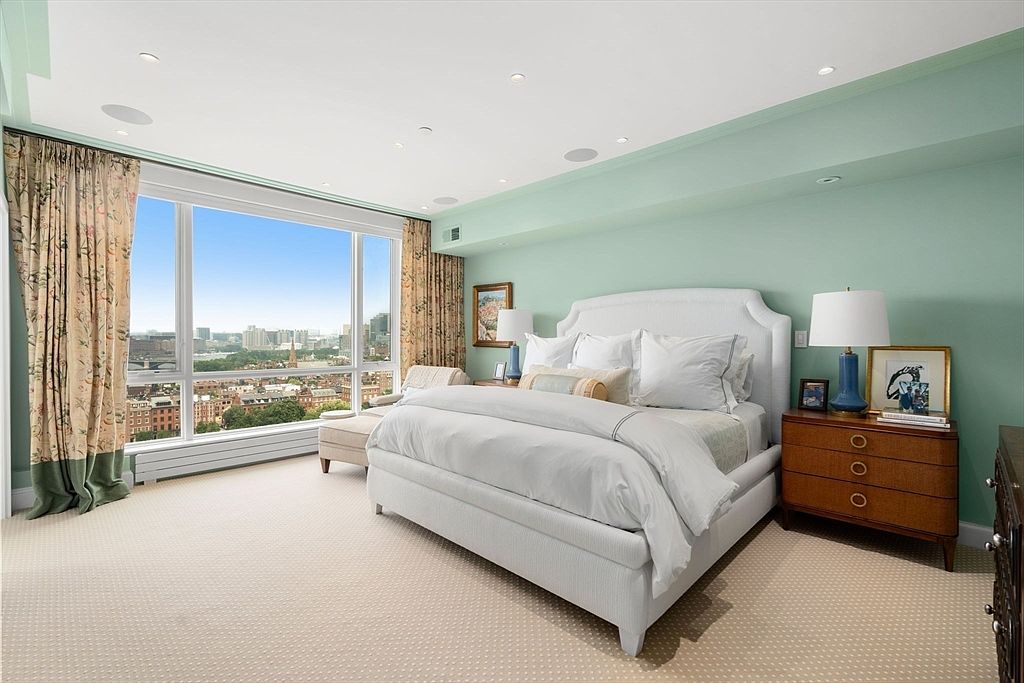 This is a primary bedroom featuring a large bed with white linens, a wooden nightstand with a lamp, and a large window with city views. The walls are painted a light green color, and the room is carpeted. The overall style is elegant and comfortable, creating a relaxing atmosphere.