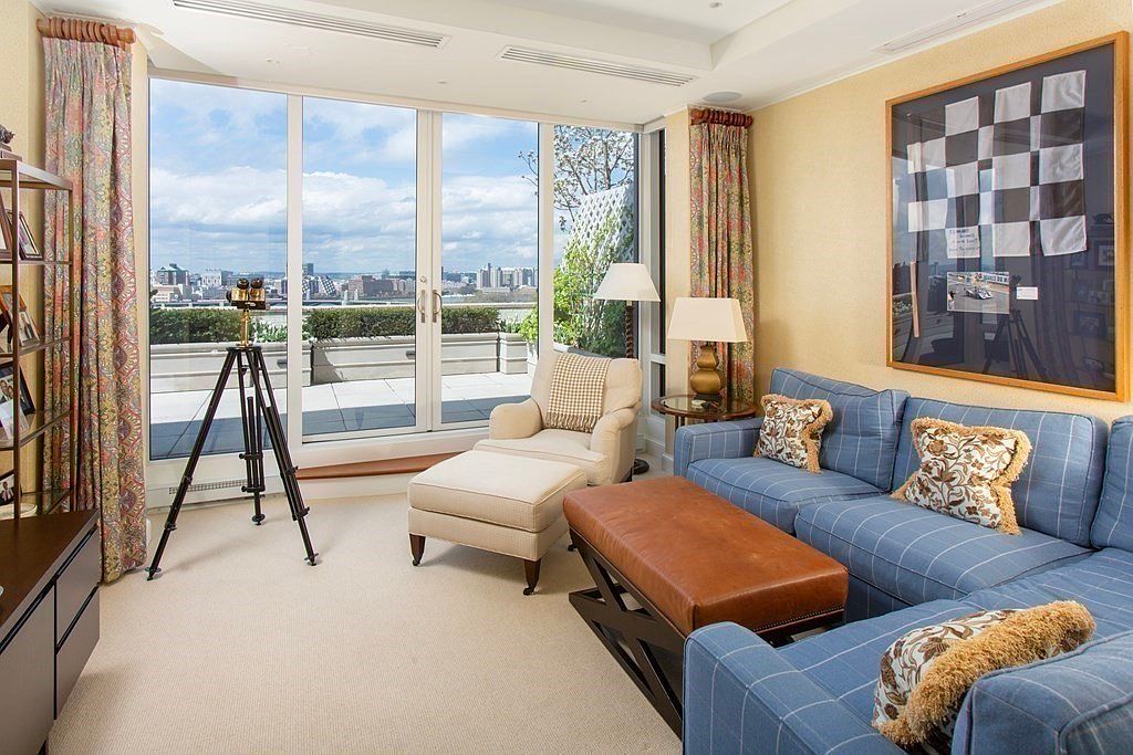 This living room features a comfortable blue plaid sofa with decorative pillows, a leather ottoman, and a beige armchair with a matching ottoman. A large window offers a stunning city view, and a telescope on a tripod suggests a focus on enjoying the scenery. The room is well-lit and decorated with a framed checkered flag artwork, creating a sophisticated and inviting atmosphere.
