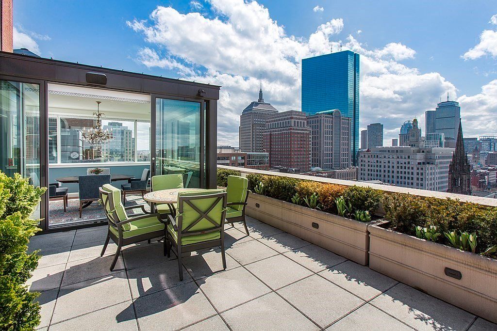 This image showcases a luxurious outdoor patio or balcony with a stunning city view. The space is furnished with a dining set featuring green cushioned chairs and a round table, complemented by a planter box filled with greenery along the edge. Sliding glass doors lead into an interior space, creating a seamless indoor-outdoor living experience.