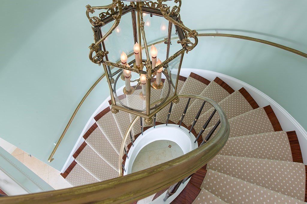 This image showcases a spiral staircase with a patterned carpet runner and dark wood trim. A decorative brass chandelier hangs above the staircase, adding a touch of elegance. The walls are painted a soft blue, and the overall impression is one of refined sophistication.