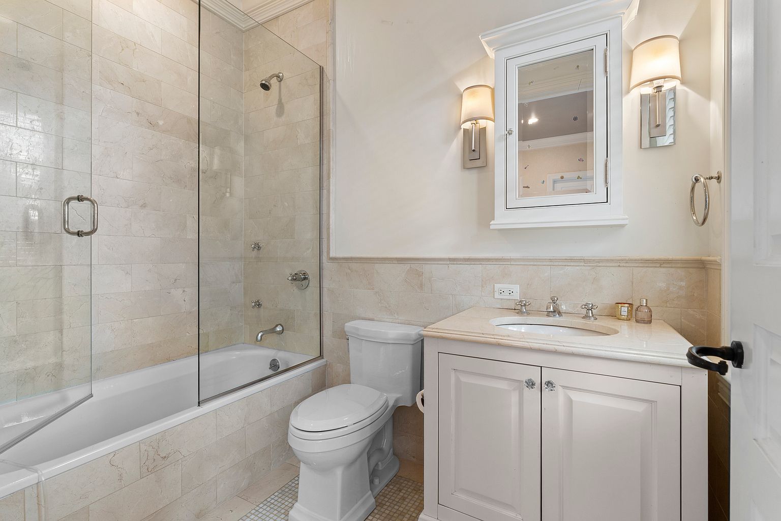 This compact, well-appointed bathroom features a neutral color palette with light-toned stone wall tiles and a matching vanity countertop. The space includes a combination tub and shower enclosed by a glass panel, a white pedestal-style toilet, and a wall-mounted medicine cabinet flanked by two elegant sconces. The overall aesthetic is clean and traditional, offering a functional and bright layout.