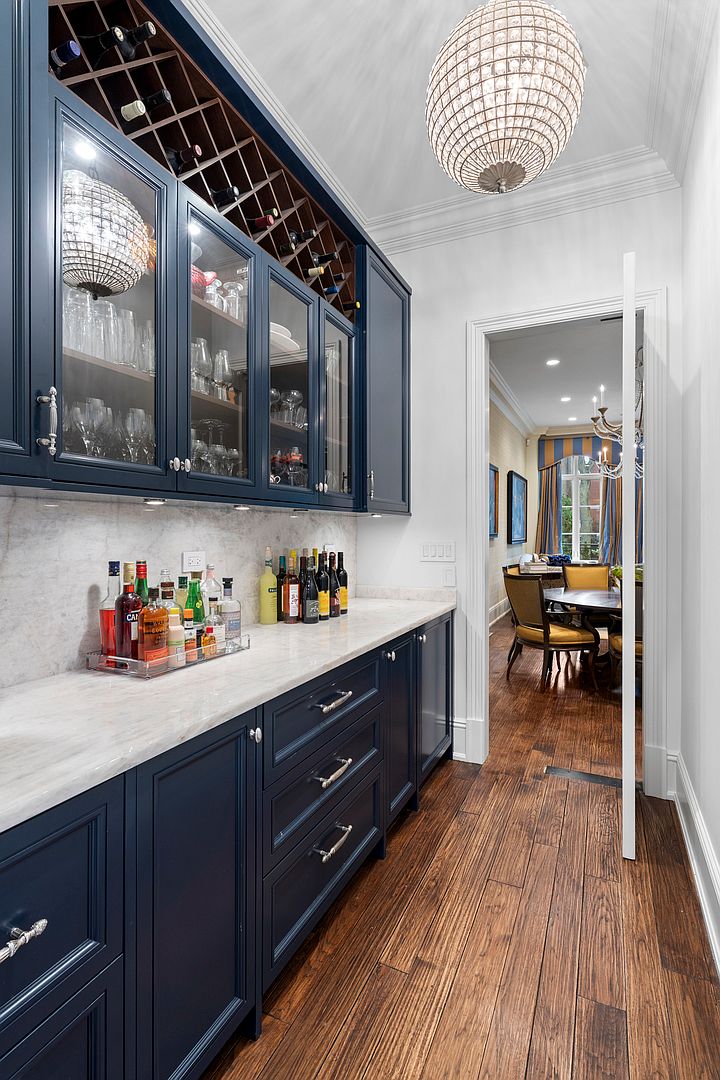 This elegant butler's pantry features deep blue cabinetry with glass-front upper cabinets and a built-in wine rack, complemented by a light-toned stone countertop and backsplash. A crystal chandelier hangs from the ceiling, adding a touch of luxury to the space, while a doorway provides a clear view into an adjacent dining room with warm wood flooring. The perspective is a wide-angle shot from the hallway, emphasizing the sophisticated transition between the service area and the formal dining space.