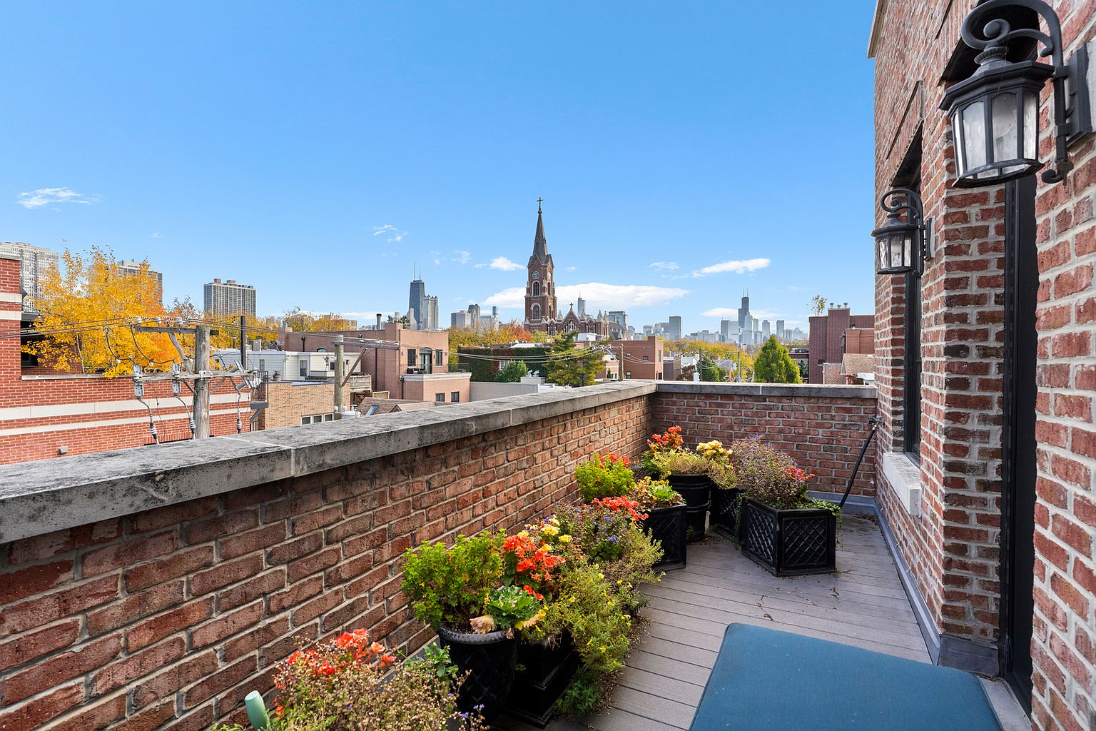This elevated rooftop terrace features a composite deck lined with brick walls and a variety of potted flowers. The space offers a panoramic view of the city skyline, including a prominent church steeple and distant skyscrapers. The perspective is from a slightly elevated vantage point, capturing the urban atmosphere and the private outdoor living area.