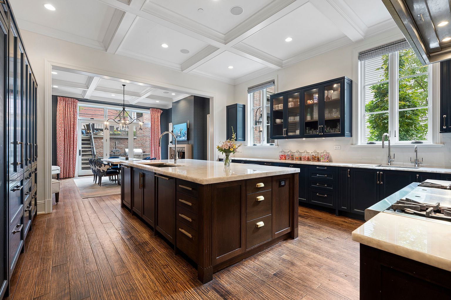This sophisticated kitchen features a large central island with a stone countertop and dark wood cabinetry, complemented by matching dark perimeter cabinets and glass-front display units. The space is anchored by rich-toned wood flooring and a coffered ceiling, creating a warm and elegant atmosphere. A wide opening leads to an adjacent dining area with views of an outdoor patio, enhancing the home's open-concept flow and natural light.