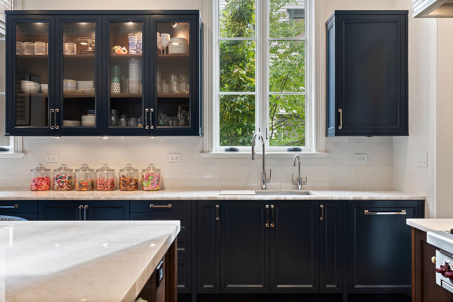 This elegant kitchen features dark navy cabinetry paired with light stone countertops and a classic white subway tile backsplash. A central window provides natural light and a view of greenery, flanked by glass-front upper cabinets and a solid-door cabinet. The space is organized with a series of glass jars filled with colorful candies on the counter, creating a playful yet sophisticated aesthetic.