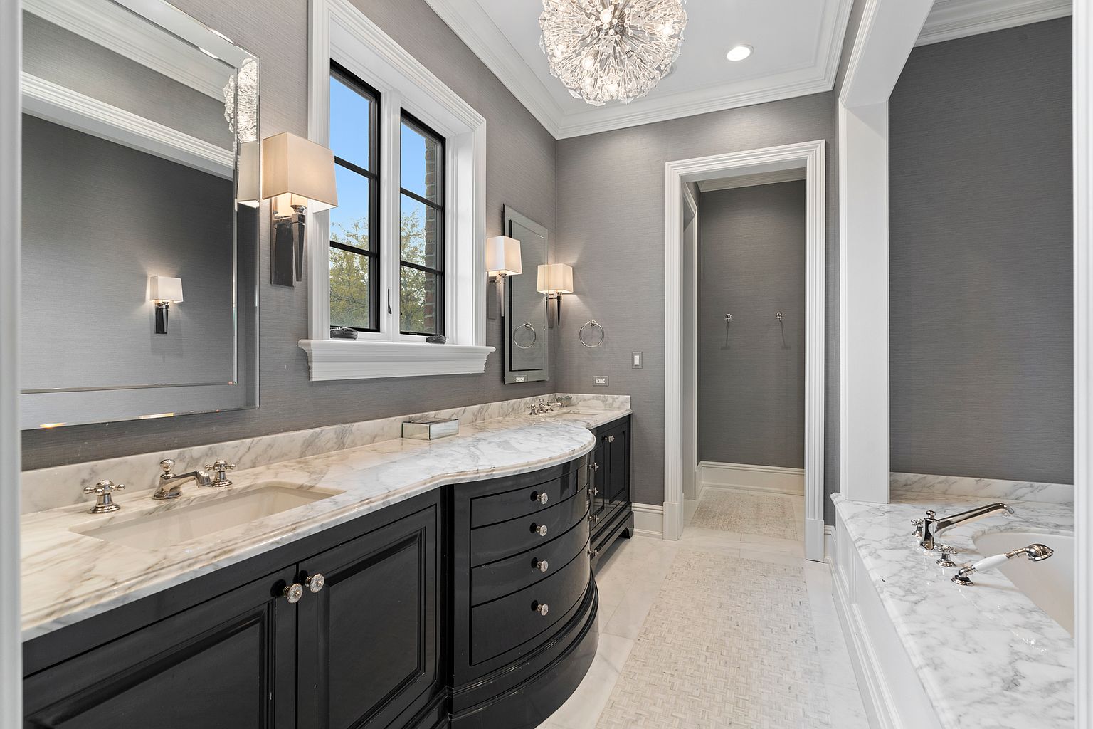 This elegant primary bathroom features a curved vanity with dark cabinetry topped by a light-veined stone countertop and dual sinks. A large window with black frames allows natural light to illuminate the space, which is accented by a crystal chandelier and wall-mounted sconces. To the right, a built-in soaking tub with stone surround completes the luxurious, high-end aesthetic of the room.