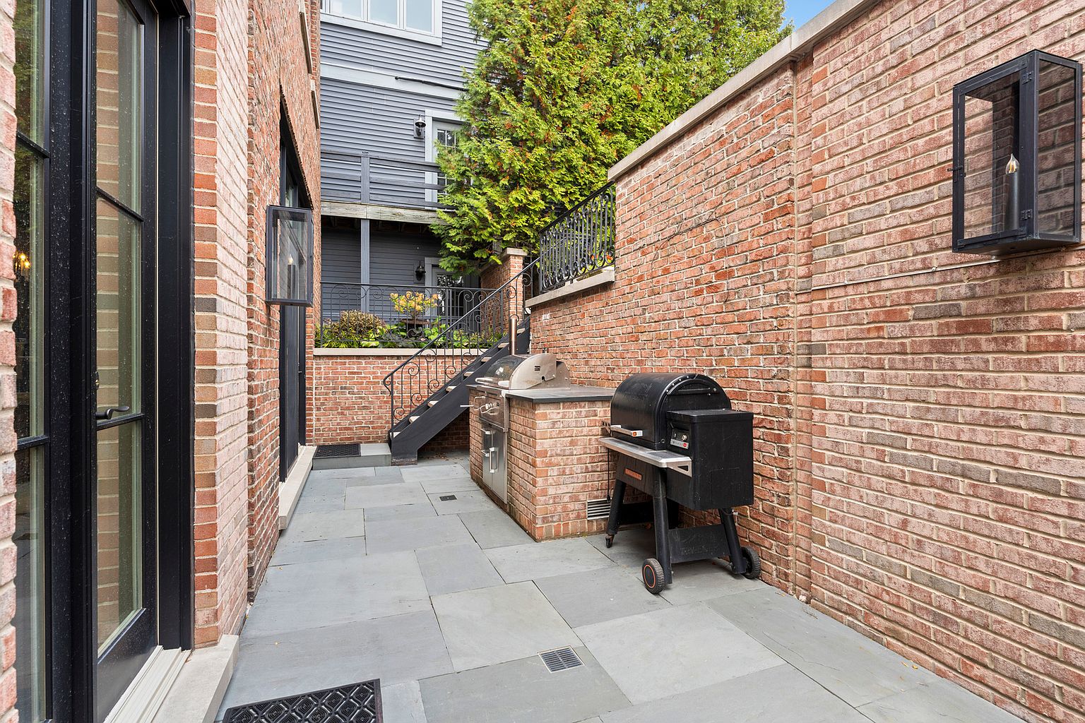 This private outdoor patio features a classic red brick aesthetic with a built-in stainless steel grill station and a standalone smoker. The space is paved with large, neutral-toned stone tiles and is framed by tall brick walls and black metal wall-mounted lanterns. A set of black metal stairs leads up to an elevated deck area, creating a multi-level outdoor living space that feels both intimate and functional.