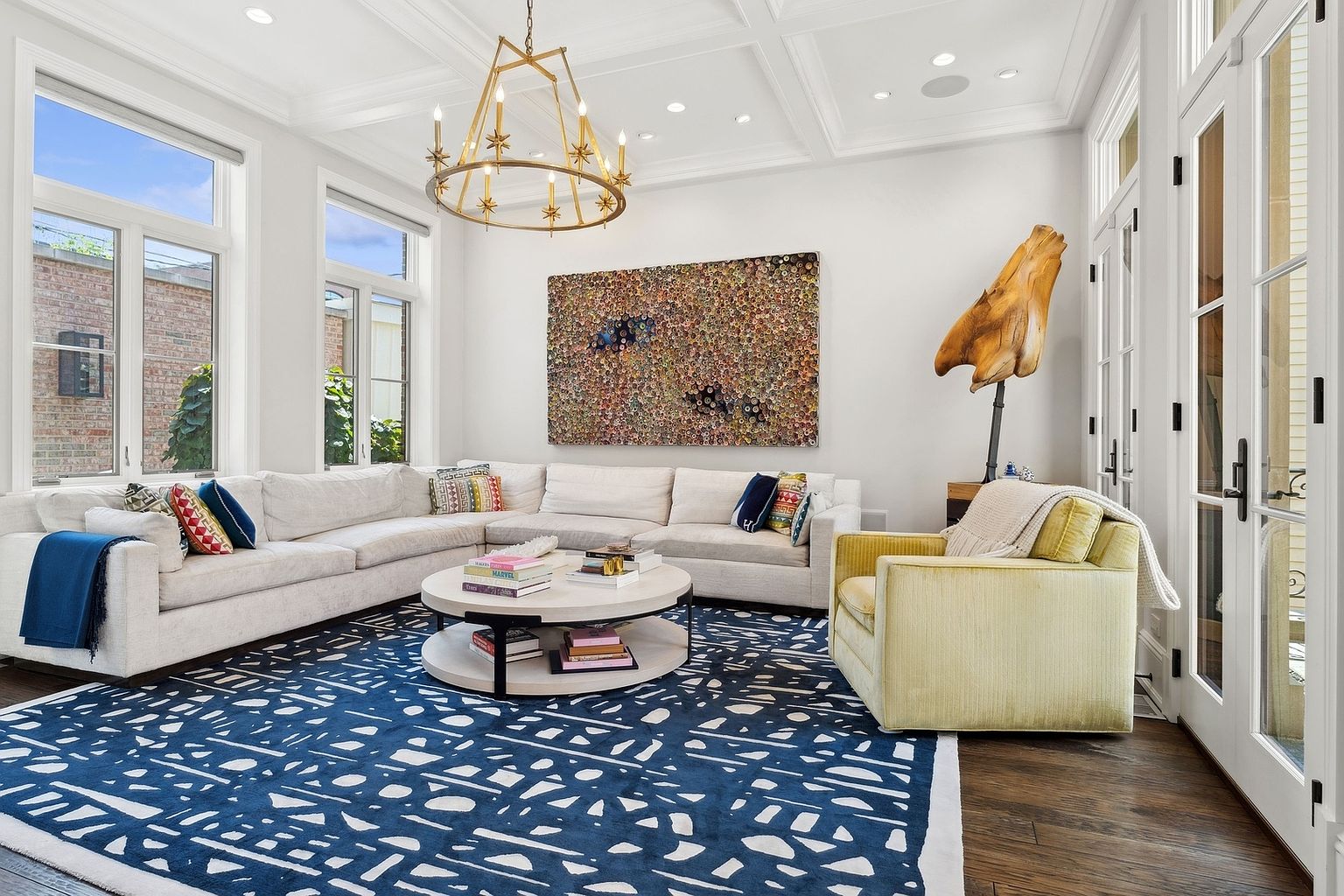 This bright and airy living room features a large, neutral-toned sectional sofa arranged around a tiered circular coffee table. A bold, navy blue patterned area rug anchors the space, while a unique sculptural art piece and a large, textured wall hanging add visual interest. The room is illuminated by a gold-toned chandelier and natural light streaming through tall windows, creating a sophisticated and welcoming atmosphere.