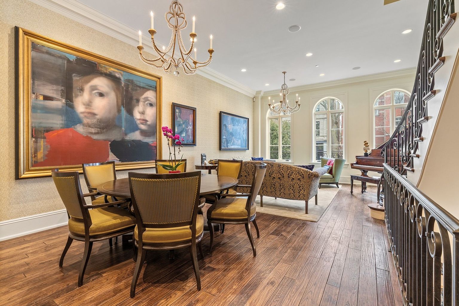 This elegant open-concept space features a formal dining area in the foreground with a round table and upholstered chairs, transitioning into a sophisticated living area. The room is adorned with large, expressive artwork, ornate chandeliers, and tall arched windows that flood the space with natural light. A grand piano sits near the staircase, which is framed by a decorative metal railing, creating a refined and luxurious atmosphere.