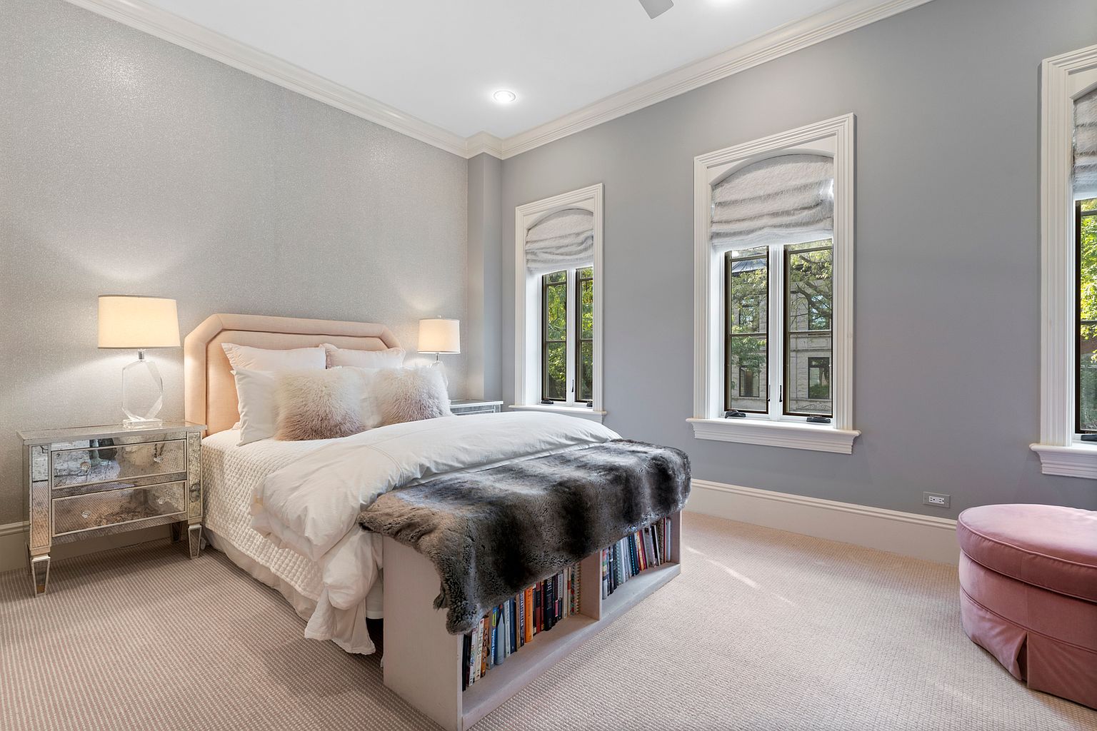 This elegant primary bedroom features a soft, neutral color palette with a textured accent wall behind a plush, light-toned headboard. The room is furnished with mirrored nightstands, a cozy bed topped with a faux-fur throw, and a built-in bookshelf at the foot of the bed. Large windows with Roman shades allow natural light to fill the space, which is completed by a soft pink ottoman and wall-to-wall carpeting.