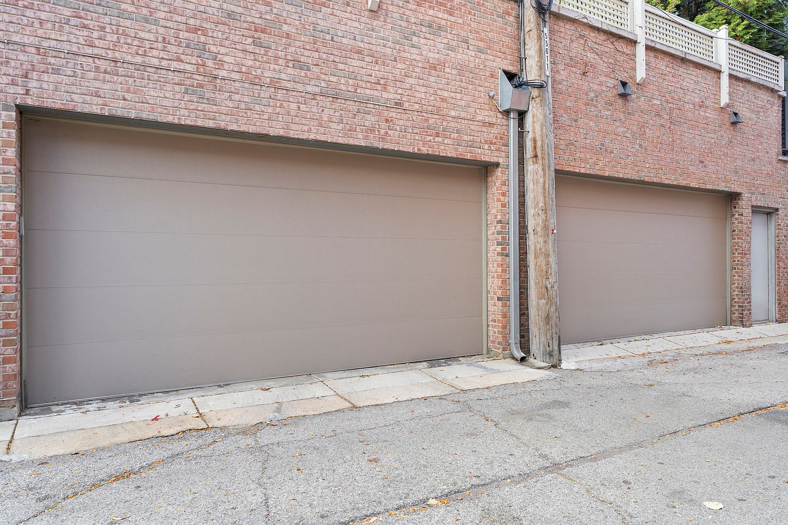 This exterior view captures a brick building featuring two large, solid-colored garage doors and a side service door. A wooden utility pole stands between the two garage bays, with a concrete alleyway running along the front. The perspective is a low-angle, eye-level shot that emphasizes the functional, utilitarian nature of the structure.