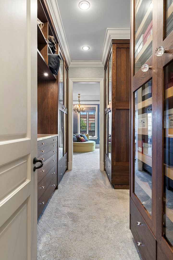 This elegant walk-in closet features rich, dark wood cabinetry with glass-fronted doors and integrated lighting, providing a sophisticated storage solution. The space is carpeted and leads directly into a bright, adjacent room visible through the doorway, which contains a round ottoman and a chandelier. The perspective is a straight-on shot looking through the closet corridor, emphasizing the organized layout and high-end finishes.