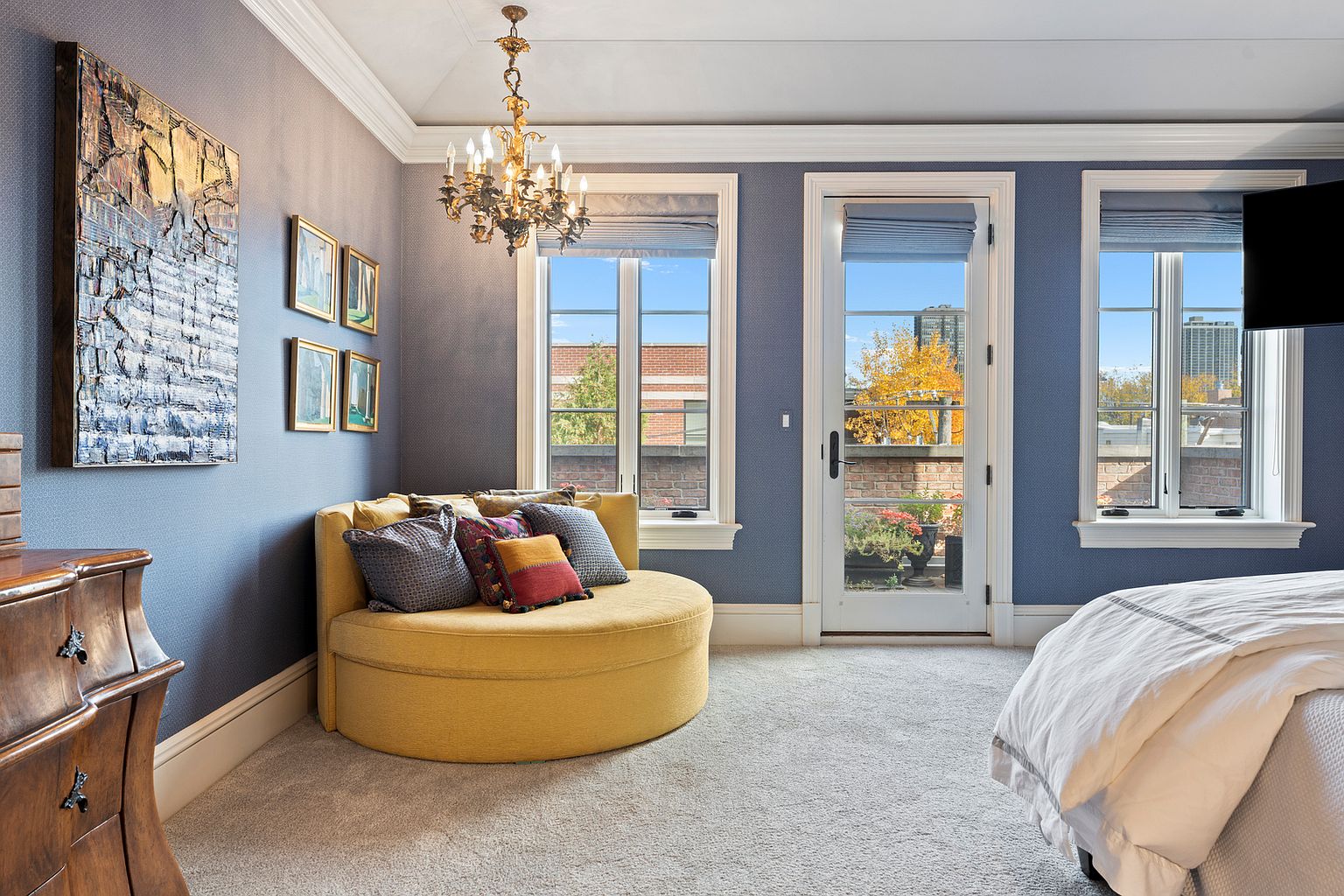 This elegant bedroom features a sophisticated blue textured wallpaper and a plush, circular yellow settee positioned near a set of windows and a glass-paned door leading to a private terrace. A classic crystal chandelier hangs from the ceiling, adding a touch of luxury, while a large piece of abstract art adorns the wall above a wooden dresser. The room is bathed in natural light, creating a serene and inviting atmosphere with a view of the surrounding neighborhood.