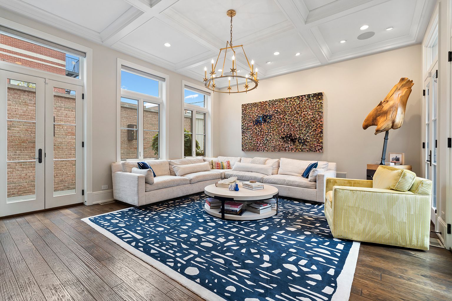 This bright and airy living room features a large, light-colored sectional sofa arranged around a circular coffee table on a bold, navy-and-white patterned area rug. A vibrant, textured wall art piece serves as a focal point above the sofa, while a unique sculptural wood piece stands on a pedestal nearby. The space is illuminated by a modern, multi-armed chandelier and benefits from natural light streaming through large windows and glass doors that open to the exterior.