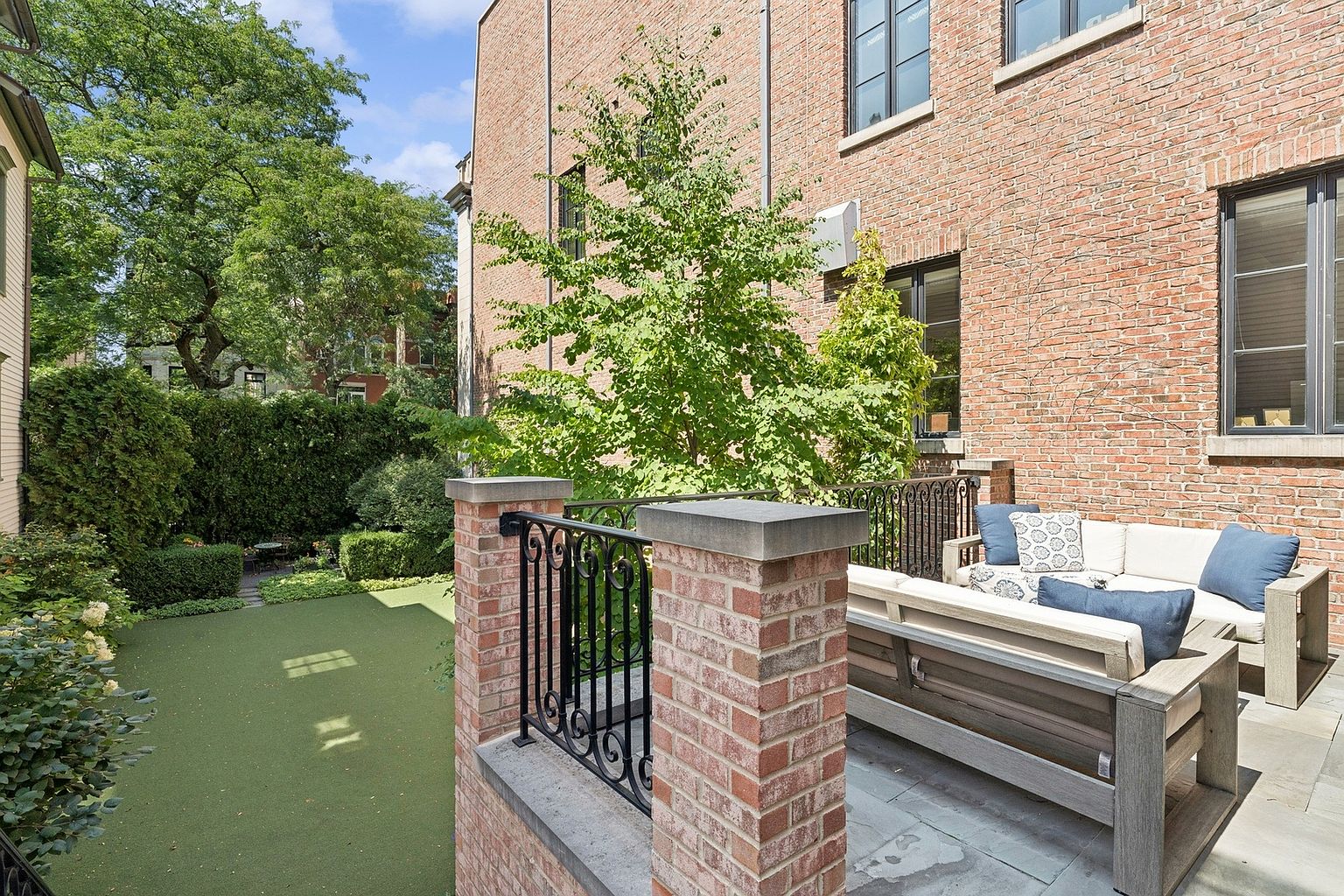 This elevated brick terrace offers a sophisticated outdoor living space, featuring a comfortable seating area with plush cushions and blue accent pillows. The patio is enclosed by a classic wrought-iron railing set between sturdy brick pillars, overlooking a manicured green lawn and lush garden foliage. The perspective captures the seamless transition from the home's brick exterior to the private backyard, creating an inviting and serene atmosphere.