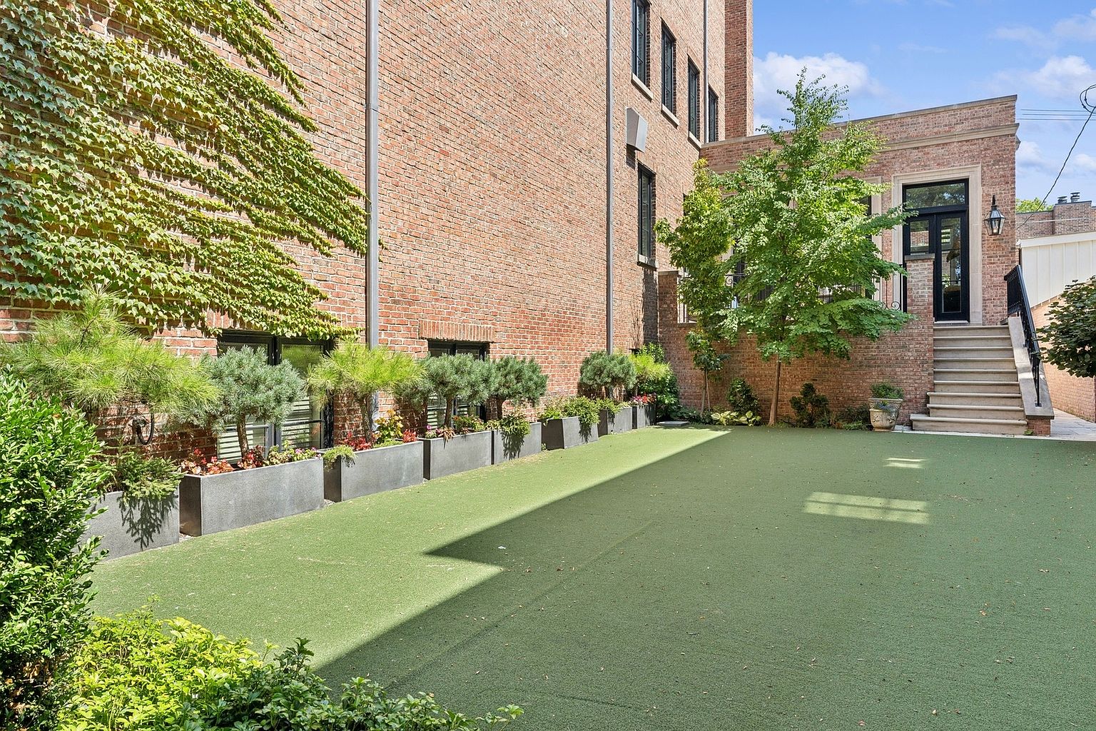 This private urban courtyard features a well-manicured artificial turf lawn bordered by a series of modern, dark-toned rectangular planters filled with lush greenery and small trees. The space is framed by a tall, ivy-covered brick wall on one side and a brick building facade with a set of stone stairs leading to an elevated entryway on the other. The perspective captures a serene, enclosed outdoor retreat that blends architectural brickwork with natural elements.