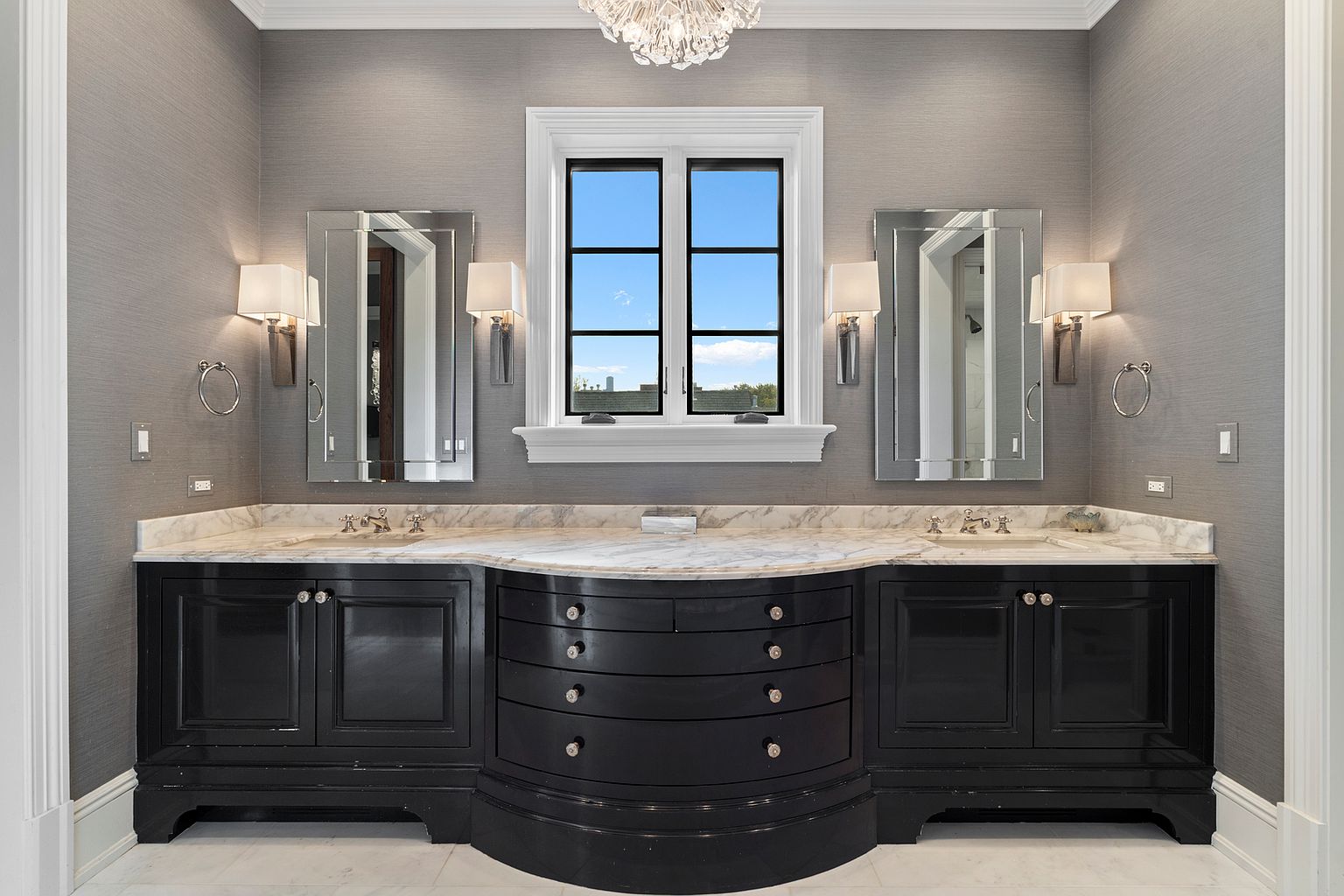 This elegant primary bathroom features a double vanity with a dark, curved central cabinet flanked by matching storage units, all topped with a light-toned stone countertop. Two rectangular mirrors are mounted on the textured gray walls, illuminated by classic wall-mounted sconces on either side. A central window with black frames provides natural light, while a crystal chandelier hangs above to complete the sophisticated, symmetrical design.