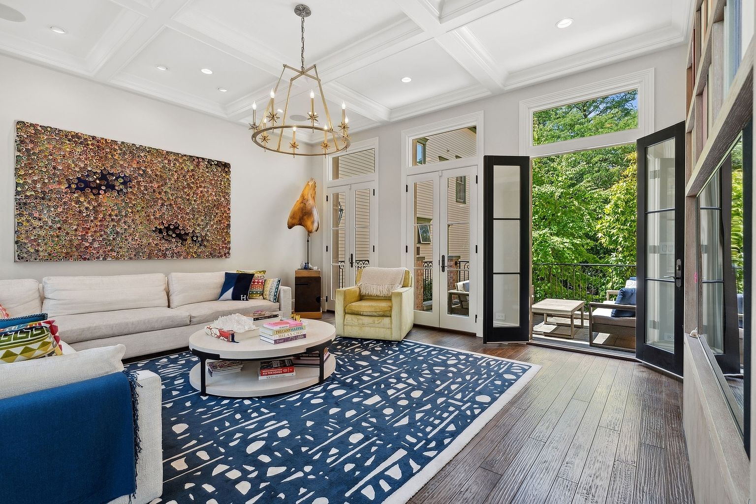 This sophisticated living room features a coffered ceiling, a large abstract art piece, and a plush white sofa paired with a patterned blue area rug. A round coffee table sits at the center, while a yellow armchair adds a pop of color near the open French doors that lead to a private balcony. The space is bright and airy, with dark wood flooring and a large chandelier providing a refined, contemporary aesthetic.