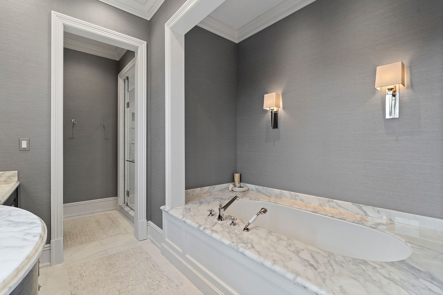 This elegant primary bathroom features a built-in soaking tub encased in a stone surround with a matching backsplash. The walls are finished in a textured gray wallcovering, complemented by two wall-mounted sconces that provide soft, ambient lighting. A doorway leads to a separate shower area, and the space is finished with light-toned patterned tile flooring, creating a sophisticated and serene atmosphere.