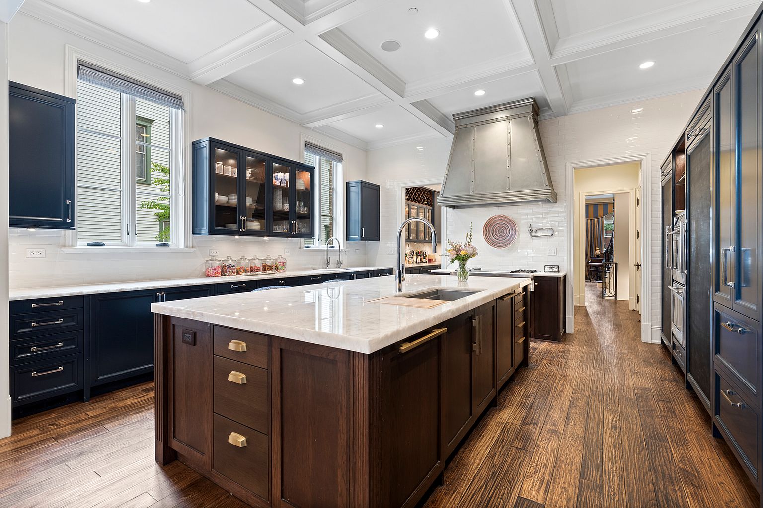 This sophisticated kitchen features a large central island with a stone countertop and dark wood cabinetry, contrasting with the perimeter's deep blue cabinets. A prominent metal range hood serves as a focal point above the stove, complemented by white subway tile backsplash and warm wood flooring. The space is brightened by recessed lighting and large windows, creating an elegant and functional culinary environment.