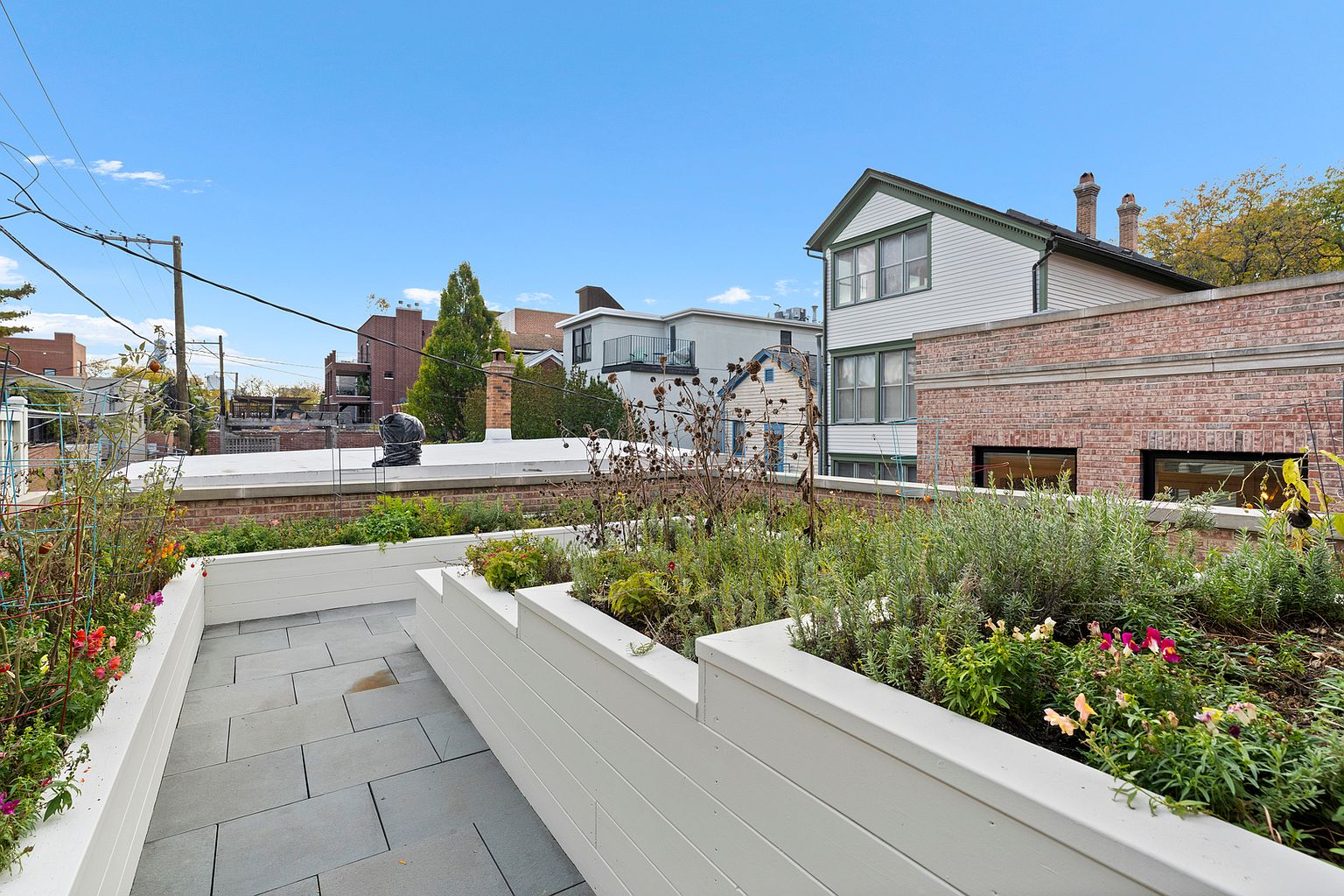 This rooftop terrace features tiered white planter boxes filled with various greenery and flowers, creating a lush urban garden atmosphere. The space is paved with large, grey stone tiles and offers an elevated perspective overlooking neighboring residential buildings. The bright, clear sky provides a clean backdrop, highlighting the well-maintained outdoor living area.