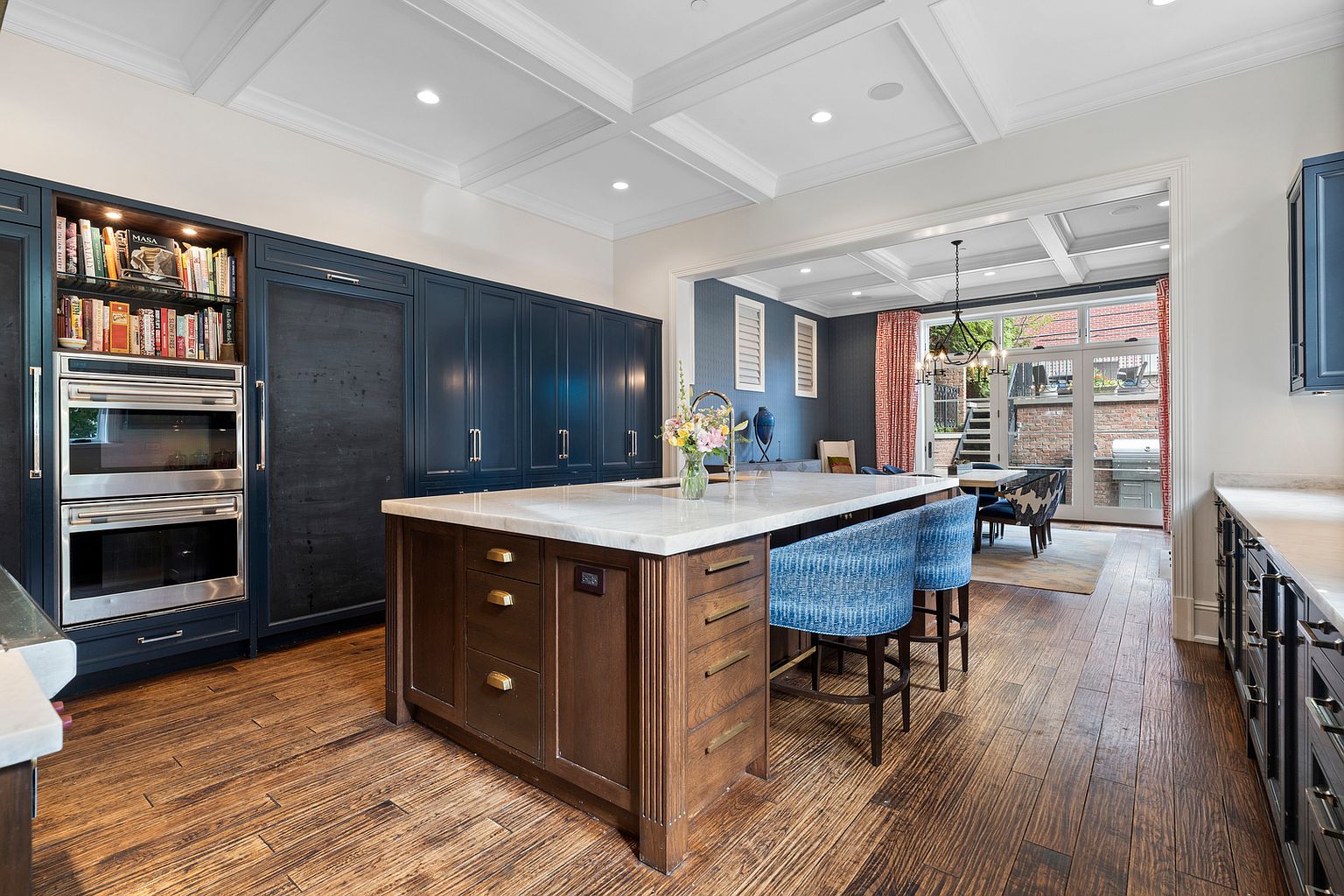 This sophisticated kitchen features dark blue cabinetry contrasted by a central island with a wood base and a light-toned stone countertop. The space is equipped with built-in stainless steel appliances and open shelving for cookbooks, while the warm wood flooring adds a natural element. A wide opening leads to an adjacent dining area with large glass doors that provide a view of the outdoor patio.