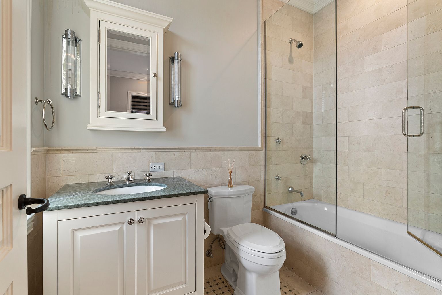 This compact, well-appointed bathroom features a white vanity with a dark stone countertop and an integrated sink, paired with a matching wall-mounted medicine cabinet. A white porcelain toilet sits beside a bathtub enclosed by a glass panel, with the walls finished in neutral-toned stone tiles. The space is illuminated by two vertical wall sconces flanking the mirror, creating a clean and functional aesthetic.