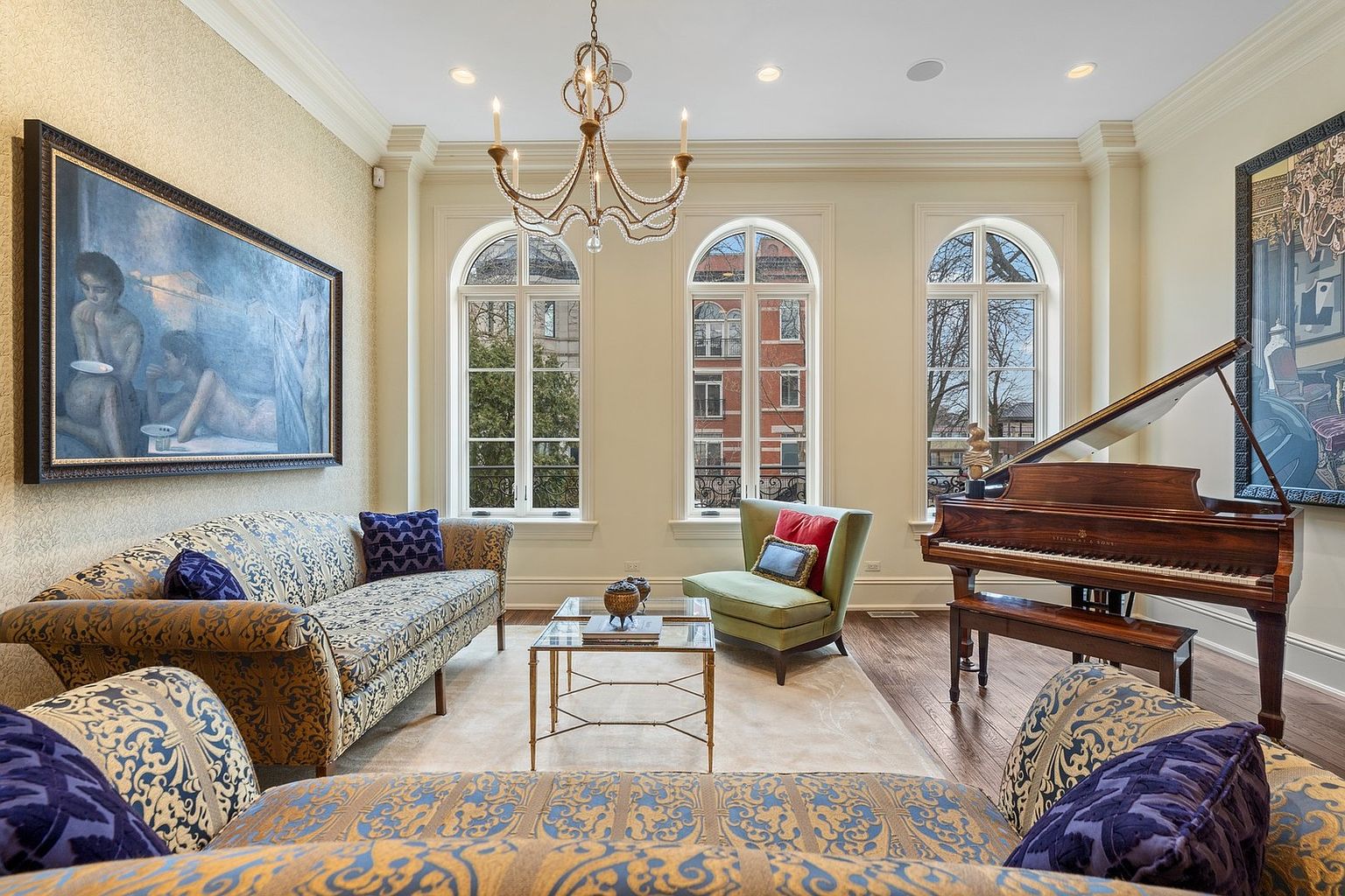 This elegant living room features a sophisticated blend of traditional and artistic elements, anchored by a grand piano and a patterned sofa. Three tall, arched windows allow ample natural light to fill the space, highlighting the dark wood flooring and the ornate chandelier suspended from the ceiling. The room is decorated with large-scale artwork on the walls, creating a refined and cultured atmosphere suitable for formal entertaining.