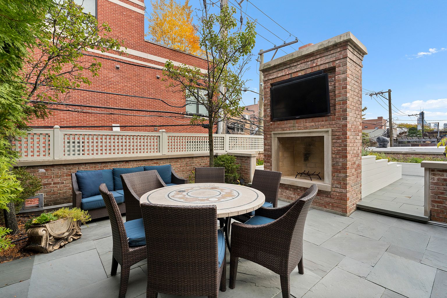 This outdoor patio space features a stone-paved floor and a central round dining table surrounded by wicker-style chairs. A prominent brick structure serves as a focal point, incorporating a built-in fireplace and a mounted television above it. The area is enclosed by a decorative lattice fence and brick walls, creating a private and inviting setting for outdoor entertaining.